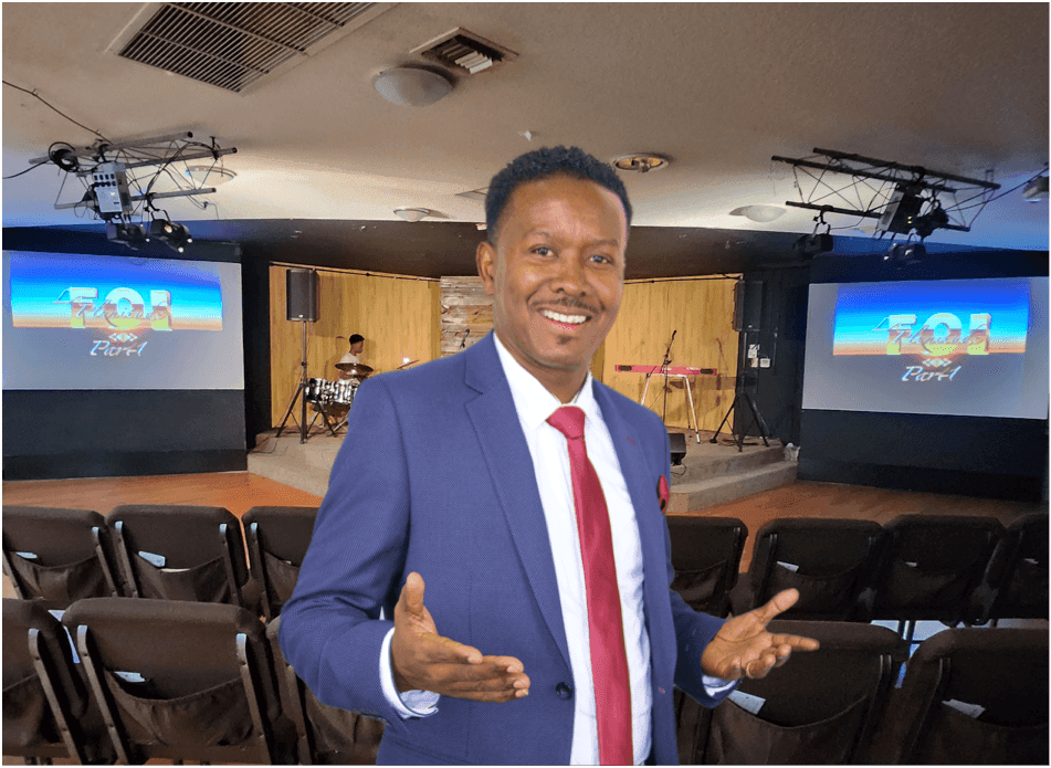 Man in a suit smiling, standing in a room with chairs and two presentation screens.